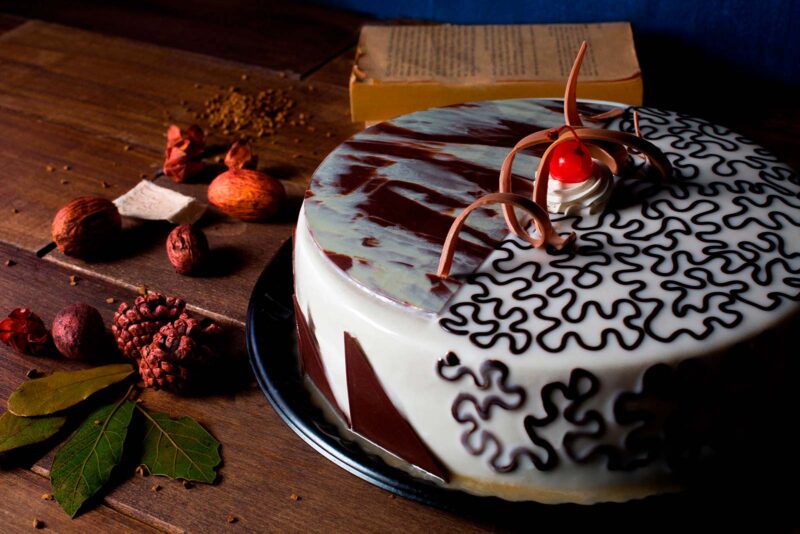 Pastel Ilusión de Lecaroz, decorado con ganache blanco, grecas de ganache oscuro, chocolate marmoleado, cereza y espiral de chocolate de leche.
