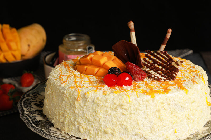Pastel Mango Go de Lecaroz, decorado con abánico y cigarros de chocolate, líneas de puré de mango, chocolate blanco rallado, crema montada, zarzamora, cereza, mango y fresa